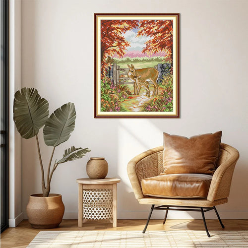 Framed artwork of a deer in a garden above a wicker chair with a plant and side table in the room.